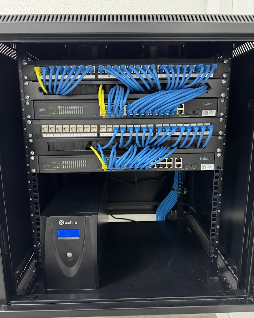 Bastidor com patch panels numerados, dois switches e uma UPS, com cabos de rede azuis organizados.