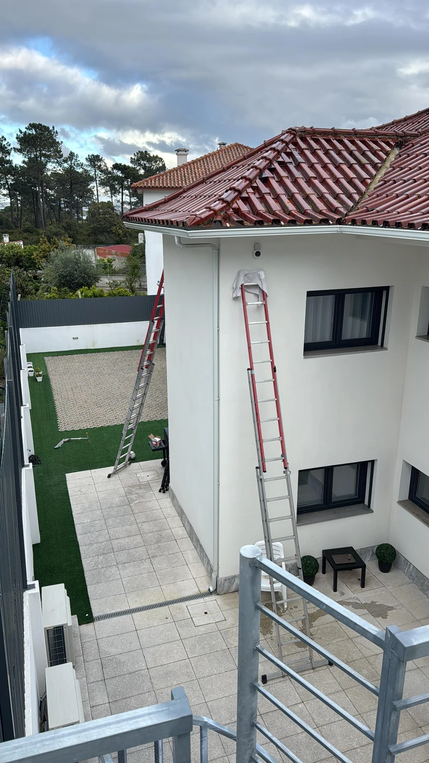 Câmara de videovigilância instalada sob o beirado de uma moradia, com escadas de trabalho encostadas.